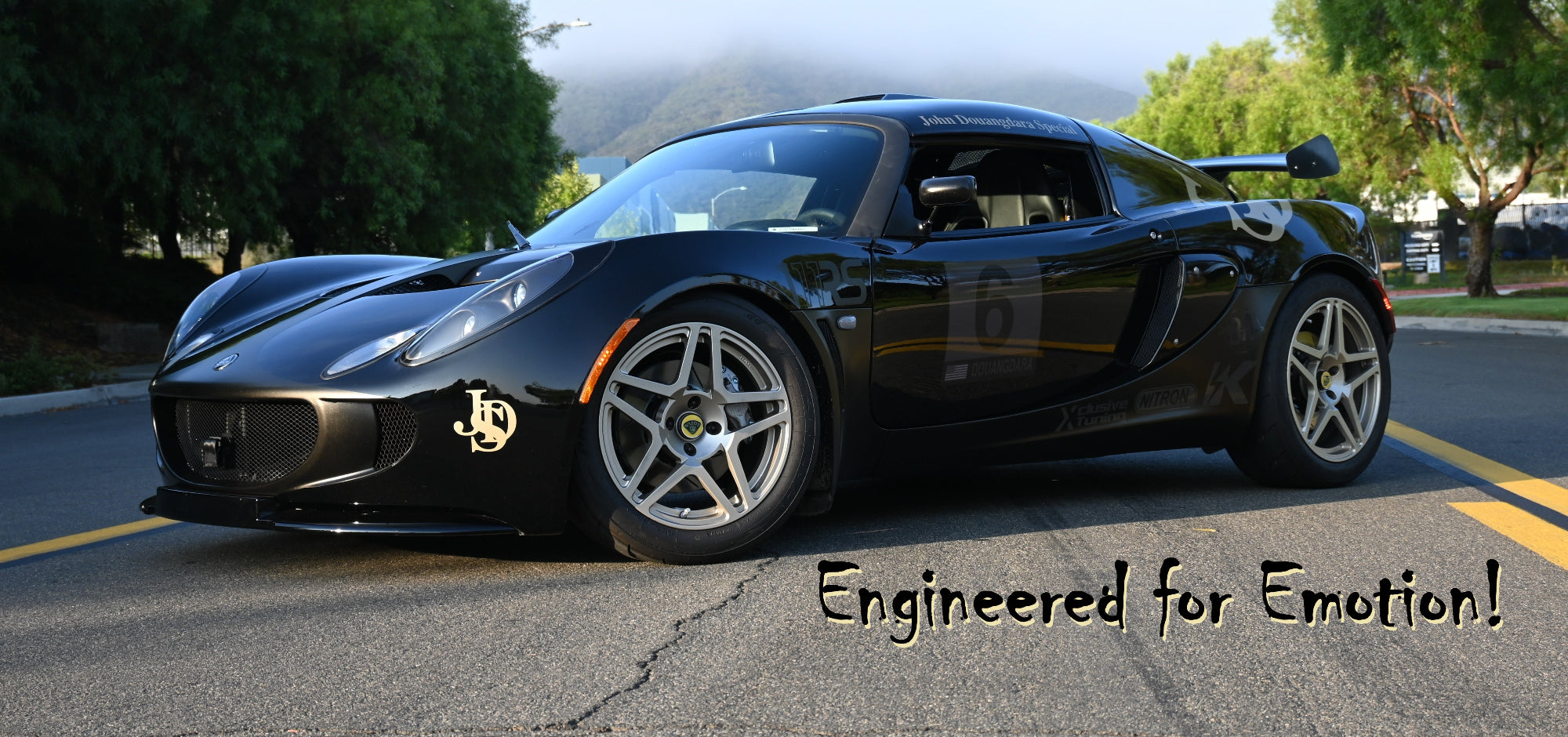 Black Lotus Exige 111RS sports car on a road with trees in the background and text 'Engineered for Emotion!'.